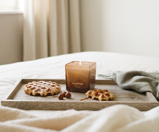 La Candela Ava&May Belgian Waffle 220 grammi accesa è appoggiata su un vassoio di legno e sopra a una tovaglia con attorno dei dolcetti waffle e delle nocciole