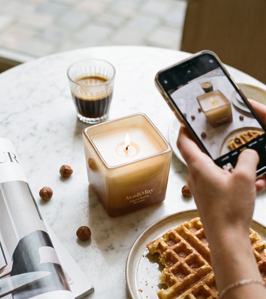 Candela Ava&May Belgian Waffle 220 grammi accesa su un tavolo di marmo con attorno un caffe espresso, un waffle belga, un giornale di interni, delle nocciole mentre viene fotografata da un telefono in mano a una persona