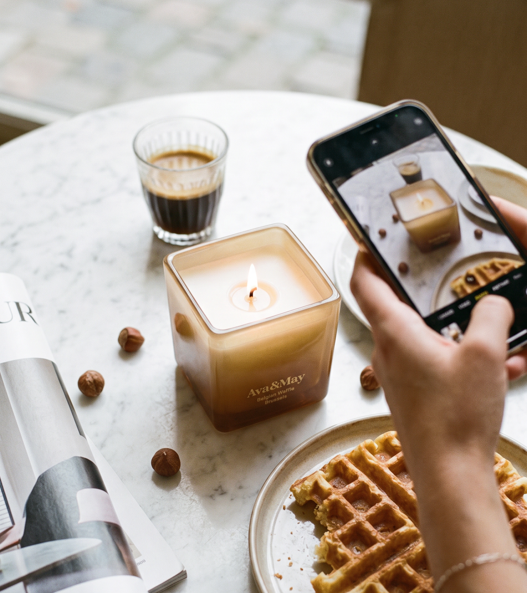 Candela Ava&May Belgian Waffle 220 grammi accesa su un tavolo di marmo con attorno un caffe espresso, un waffle belga, un giornale di interni, delle nocciole mentre viene fotografata da un telefono in mano a una persona