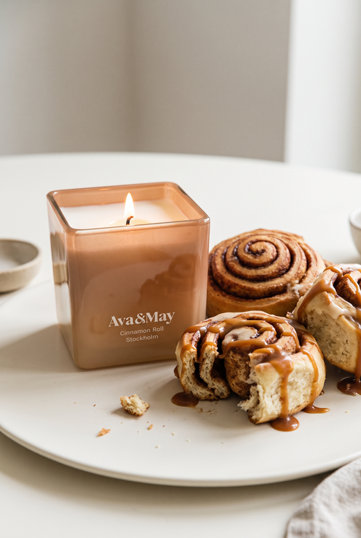 La Candela Cinnamon Roll 220 grammi di Ava&May accesa un piattino di ceramica con vicino dei dolcetti alla cannella speziati e ricoperti di crema al caramello