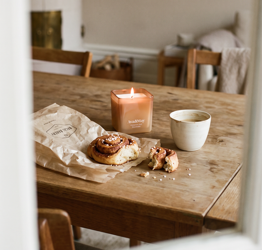 Candela Cinnamon Roll 220 grammi di Ava&May accesa su un tavolo di legno di una sala da pranzo con vicino una tazza di caffè e un dolcetto alla cannella di un panificio