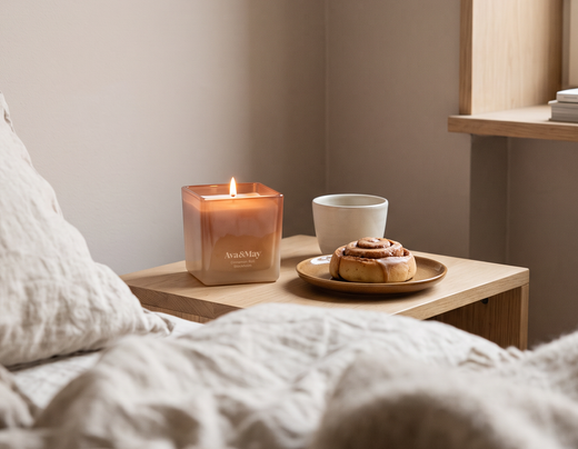 La Candela Cinnamon Roll 220 grammi di Ava&May accesa su un comodino di legno vicino a un letto con lenzuola beige. Vicino ha un dolcetto panificato alla cannella e una tazza bianca