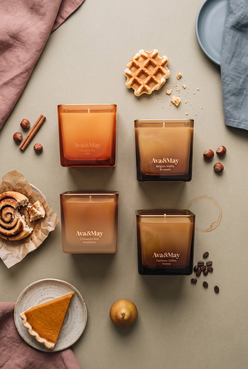 Set di candele Sweet Sensations di Ava&May: le candele ai profumi di Belgian Waffle 220 grammi, Viennese Coffee 220 grammi, Pumpkin pie 220 grammi e Cinnamon Roll 220 grammi sono sdraiate su un tavolo verde e vicino hanno una torta alla zucca, un dolcetto alla cannella, della cannella, un waffle belga, nocciole e chicchi di caffe