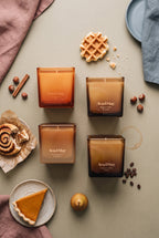 Set di candele Sweet Sensations di Ava&May: le candele ai profumi di Belgian Waffle 220 grammi, Viennese Coffee 220 grammi, Pumpkin pie 220 grammi e Cinnamon Roll 220 grammi sono sdraiate su un tavolo verde e vicino hanno una torta alla zucca, un dolcetto alla cannella, della cannella, un waffle belga, nocciole e chicchi di caffe