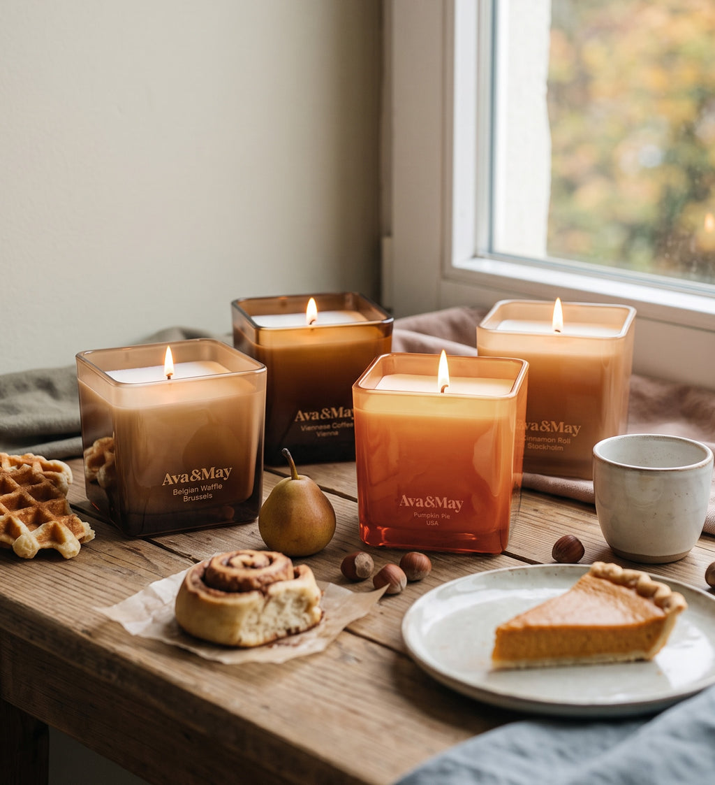Set di candele Sweet Sensations di Ava&May: candele ai profumi di Belgian Waffle 220 grammi, Viennese Coffee 220 grammi, Pumpkin pie 220 grammi e Cinnamon Roll 220 grammi, accese su un tavolo di legno con waffle, torta di zucca e rotoli alla cannella