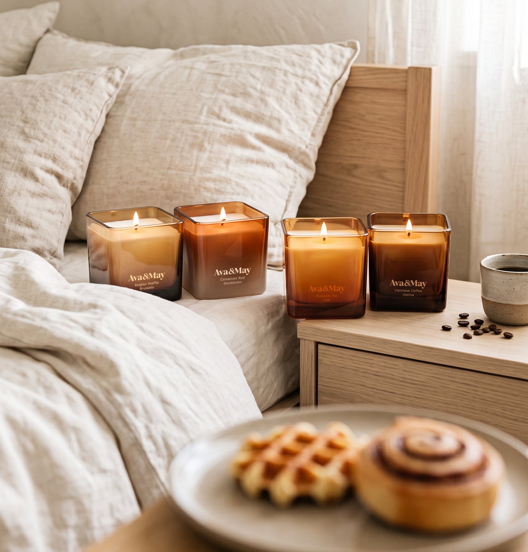 le candele Belgian waffle 220 grammi e Cinnamon Roll 220 grammi sono accese e appoggiate sul letto, mentre le candele Viennese Coffee 220 grammi e Pumpkin Pie 220 grammi sono sul comodino vicino a una tazza di caffe e dei chicchi di caffe. in primo piano c‘è un piattino con dei dolcetti